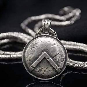 May include: A silver-toned pendant necklace featuring a shield design with a chevron and a textured surface. The pendant hangs from a detailed chain, creating a classic and historical aesthetic. The necklace is displayed against a dark background.
