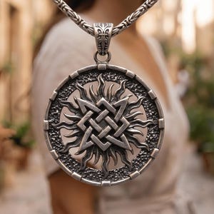 May include: A silver pendant necklace featuring a detailed circular design with a central knot pattern and sun-like rays. The pendant hangs from a silver chain, showcasing intricate metalwork. The necklace is a piece of jewelry.