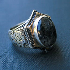 May include: A silver ring with a large, oval, dark gray stone. The ring has a gold-colored bezel and intricate floral and filigree detailing on the band. The stone is set in a gold-colored bezel with a silver border.