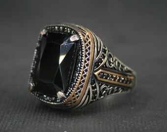 May include: A silver ring with a large, rectangular black gemstone. The ring features intricate filigree and small black stones around the main stone and on the band. Accents of gold-colored metal are visible.