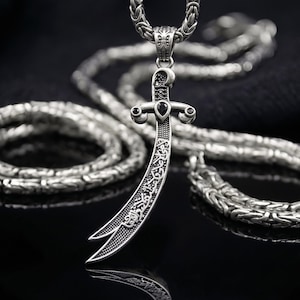 May include: A silver pendant necklace featuring a curved sword design with intricate patterns and a black gemstone. The pendant hangs from a thick, textured chain. The background is black, highlighting the necklace's details.