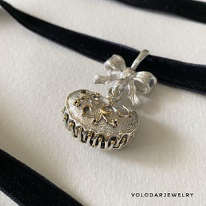 Puede incluir: Un collar gargantilla plateado con una cinta de terciopelo negro. El colgante tiene forma ovalada con detalles intrincados y detalles dorados, suspendido de un lazo plateado. El collar se muestra sobre una superficie blanca.