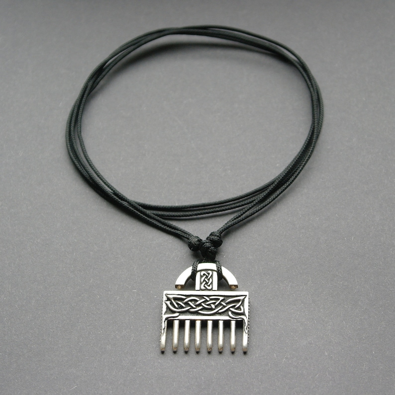 Puede incluir: Un collar con un colgante de plata con un dise&ntilde;o de nudo celta y una parte inferior en forma de peine. El collar est&aacute; en un cord&oacute;n negro.