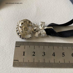 May include: A silver choker necklace with a decorative bow and a detailed clasp. The clasp has an ornate design with gold accents. The black velvet band is attached to the clasp and bow. The ruler shows the clasp is approximately 1 inch in length.