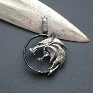 May include: A silver-colored pendant featuring a wolf's head within a circular frame. The wolf is snarling, with detailed fur and teeth. The pendant has a loop for a chain and is set against a dark background with a light-colored object.