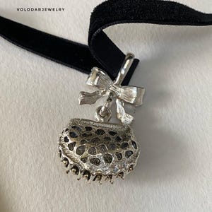 May include: A silver-toned pendant necklace with a black velvet ribbon. The pendant features a bow and a small, openwork purse-shaped charm. The jewelry is from VOLODARJEWELRY.