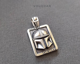 May include: A rectangular silver-toned pendant featuring a raised helmet design in the center. The text THIS IS THE WAY is embossed along the top and bottom edges of the frame. A metal bail is attached to the top for hanging on a chain.