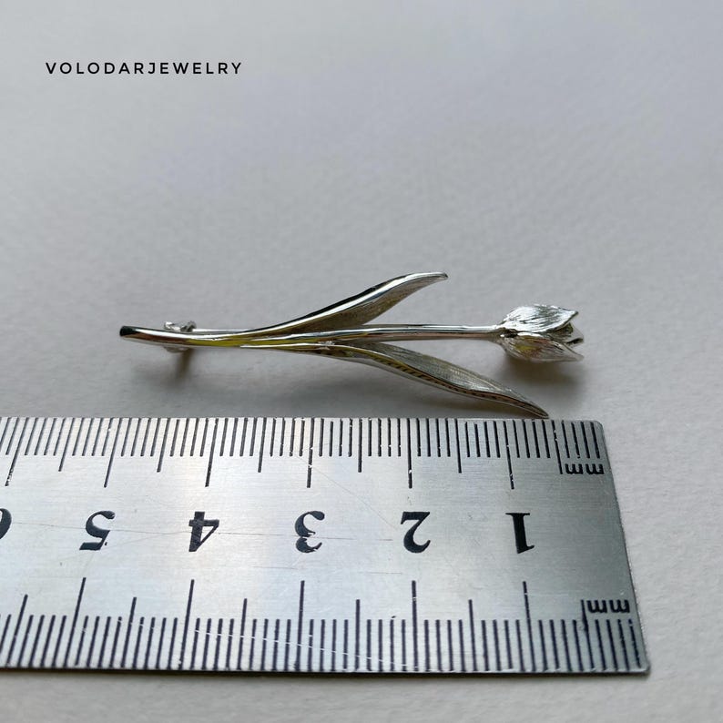 May include: A silver tulip brooch, approximately 5 cm in length. The brooch features detailed leaves and a delicate flower head. The jewelry is set against a neutral background. The text "VOLODARJEWELRY" is visible in the upper left corner.