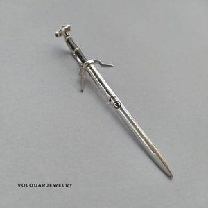 Puede incluir: Un colgante de plata en forma de espada con mango negro. La espada tiene un diseño de cabeza de lobo en la empuñadura y la inscripción "The Witcher" en la hoja.