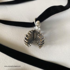 May include: A silver-toned pendant necklace featuring a detailed rib cage design. The pendant is suspended from a black velvet choker. The necklace has a gothic or anatomical theme, suitable for alternative fashion.