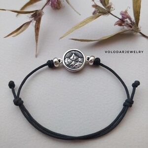Könnte beinhalten: Ein schwarzes Kordelarmband mit einem silbernen Anhänger, der ein Gebirgsmassiv zeigt. Der Anhänger ist rund und hat eine strukturierte Oberfläche. Das Armband ist an beiden Enden mit einem Knoten befestigt.