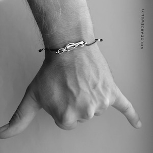 Könnte beinhalten: Ein schwarzes Kordelarmband mit einem silbernen Anhänger in Form eines Knotens. Das Armband befindet sich am Handgelenk einer Person.
