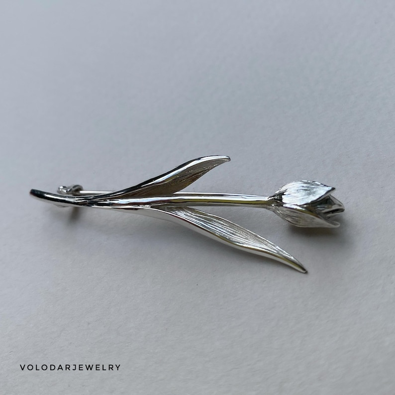 May include: A silver tulip brooch with detailed petals and leaves. The brooch is a realistic depiction of a tulip flower, with a pin clasp on the back. The jewelry is a floral accessory.