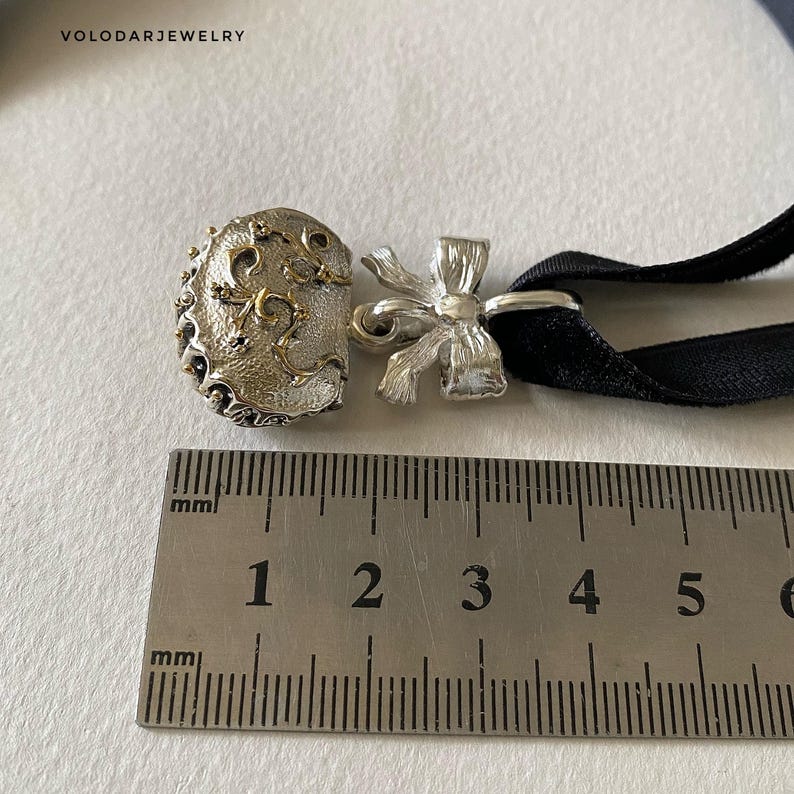 May include: A silver choker necklace with a decorative bow and a detailed clasp. The clasp has an ornate design with gold accents. The black ribbon measures approximately 0.5 inches wide. The necklace is displayed on a white surface.