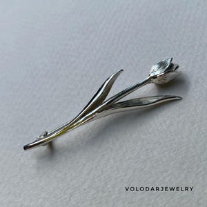 May include: A silver tulip brooch with intricate petal and leaf detailing. The brooch depicts a realistic tulip flower, featuring a long stem and two leaves. The jewellery is likely made of sterling silver.