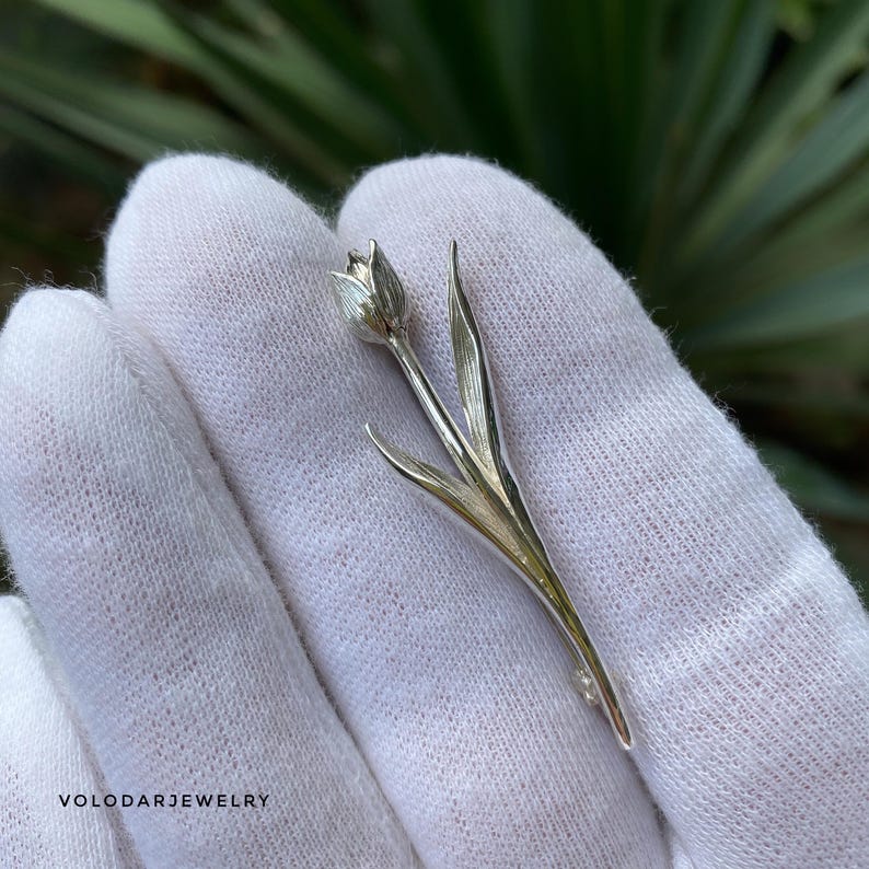 May include: A silver tulip brooch with detailed leaves and a delicate flower bud. The brooch is displayed on a white surface. The jewelry is from VOLODARJEWELRY.