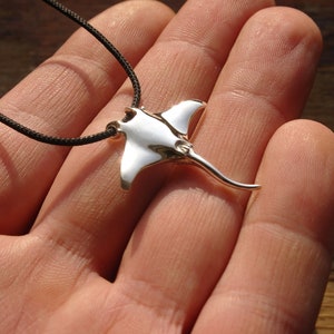 Puede incluir: Un colgante de manta raya de color plateado en un collar de cordón negro. El colgante es detallado y tiene una forma realista.