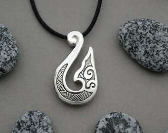 May include: A silver pendant necklace with a detailed Maori fish hook design. The pendant is hanging from a black cord.