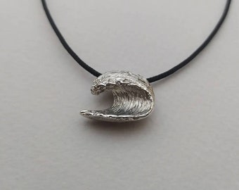 Può includere: Una collana con un ciondolo a forma di onda argentata su un cordoncino nero. Il ciondolo è un'onda tridimensionale testurizzata e dettagliata.