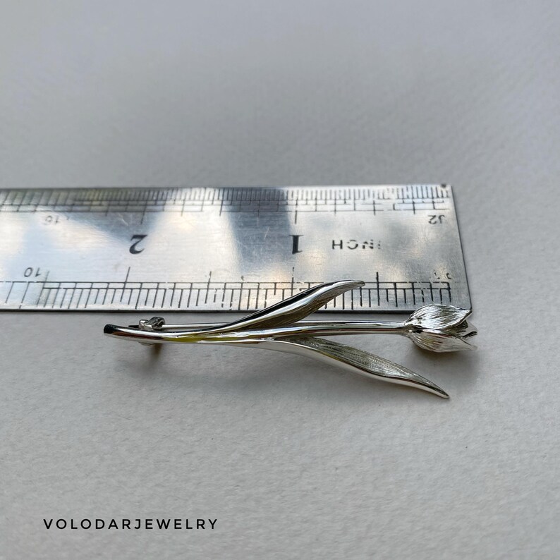 May include: A silver tulip brooch with detailed leaves and petals. The brooch is approximately 5 cm long, as measured by a ruler. The brooch is a floral accessory, perfect for adding a touch of nature-inspired elegance to any outfit.