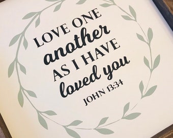 May include: A framed sign with the text "LOVE ONE another AS I HAVE loved you JOHN 13:34". The text is in black script and is surrounded by a light green leaf wreath on a white background. The frame is made of dark brown wood.
