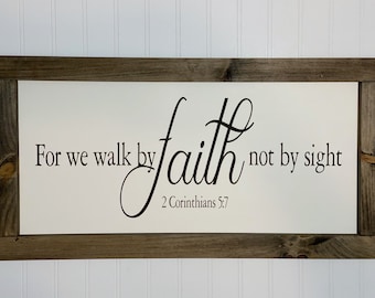 May include: A rectangular wooden sign with a white background and a dark brown frame. The sign features the text "For we walk by faith not by sight" and "2 Corinthians 5:7" in black script.
