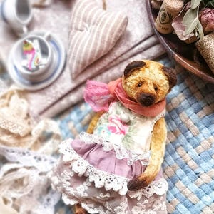 Könnte beinhalten: Ein handgefertigter Teddybär mit einem geblümten Kleid mit Spitzenbesatz und einem rosa Schal. Der Bär hat braunes Fell und dunkelbraune Ohren. Das Kleid ist helllila. Der Hintergrund umfasst eine herzförmige Dekoration und eine kleine Teetasse.