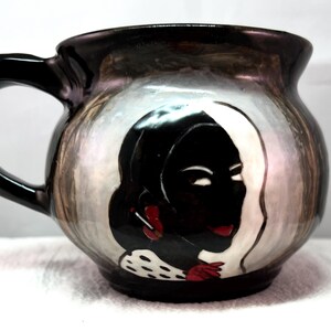 Puede incluir: Una taza de cerámica con asa y borde negros, que presenta un retrato pintado de una mujer con cabello negro y detalles rojos. La taza tiene un acabado nacarado con un fondo blanco y marrón. La taza es probablemente hecha a mano.