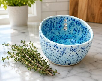 Puede incluir: Un cuenco de cerámica con base azul claro y un patrón de motas azul oscuro. El cuenco presenta tres pequeños orificios circulares alineados verticalmente cerca del borde superior. Una rama de tomillo verde fresco se sitúa frente al cuenco.