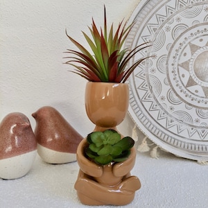 May include: A ceramic planter shaped like a person sitting in a meditative pose, holding a small succulent plant in their lap. The planter is a light brown colour and has a smooth, glossy finish. The succulent is green and has a variety of textures.