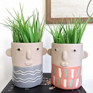 May include: Two ceramic planters shaped like faces with green grass growing out of the top. The planter on the left has a grey and white wavy pattern and the planter on the right has a pink and white striped pattern.