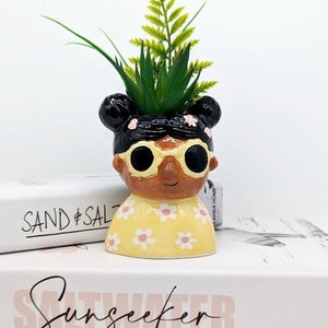 May include: A ceramic planter shaped like a person with black hair, brown skin, and yellow sunglasses. The planter is wearing a yellow shirt with pink flowers and has a green plant growing out of the top. The planter is sitting on top of two books, one titled "SAND & SALT" and the other titled "Sunseeker".