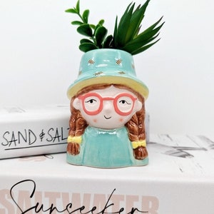 Puede incluir: Un macetero de cerámica con forma de niña con un sombrero azul, gafas rojas y trenzas marrones. El macetero tiene un diseño de patrón azul claro y blanco. El macetero contiene una pequeña planta verde.
