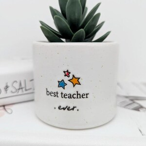 May include: White ceramic planter with a succulent plant. The planter has a speckled glaze and says "best teacher . ever." with three colorful stars.