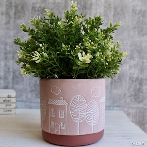 May include: A pink ceramic planter with a white design of a house and trees. The planter is filled with a green leafy plant.