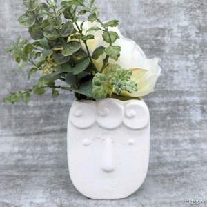 Puede incluir: Maceta de cerámica blanca con forma de cara y un diseño caprichoso. La maceta tiene una superficie texturizada y presenta una pequeña nariz y ojos. La maceta está llena de un ramo de flores blancas y follaje verde.