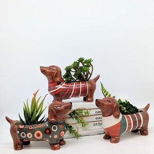 May include: Three ceramic dachshund planters, each with a brown body and a different patterned coat in red, green, and grey. Each planter holds a small succulent plant. The planters are arranged on a white surface.