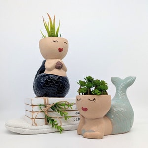 May include: Two mermaid planter pots, one with a black tail and one with a light blue tail. Both planters have a light brown face with closed eyes and a red heart-shaped mouth. The black mermaid planter has a small green succulent plant inside. The light blue mermaid planter has a small green succulent plant inside.