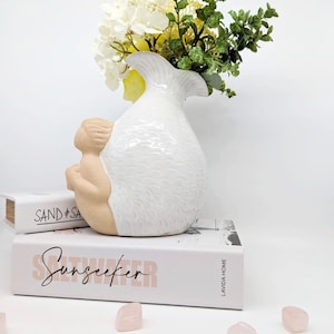 May include: White ceramic vase shaped like a mermaid with a tail and a human-like figure. The vase is filled with white and yellow flowers and green leaves. The vase is sitting on top of two books, one with the text "SAND & SA" and the other with the text "Sunseeker" and "LAVIDA HOME".