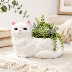 Puede incluir: Una maceta de cerámica blanca con forma de gato de pelo largo, con ojos azules y nariz rosa. La maceta contiene un conjunto de plantas suculentas verdes, con algunas hojas que cuelgan sobre el costado del lomo del gato.