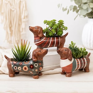 May include: Three ceramic dachshund-shaped planters with glossy brown finishes. Each dog wears a different patterned jumper: one with red and brown stripes, one with multicoloured circles on a grey background, and one with green, red, and white stripes.