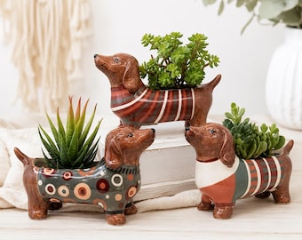 May include: Three ceramic dachshund-shaped planters with glossy brown finishes. Each dog wears a different patterned sweater: one with red and brown stripes, one with multicolored circles on a gray background, and one with green, red, and white stripes.
