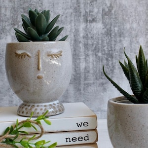 Pode incluir: Dois vasos de cerâmica com um esmalte branco e um acabamento salpicado. O vaso à esquerda tem um rosto com os olhos fechados e um nariz. O vaso à direita está vazio. Ambos os vasos estão sentados em uma pilha de livros brancos com as palavras "we need" escritas no livro superior.