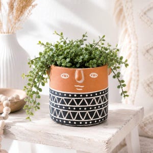 Puede incluir: Una maceta cilíndrica de terracota con un rostro estilizado de líneas blancas en la parte superior y una base negra decorada con triángulos y puntos geométricos blancos, que contiene una planta verde colgante de hojas pequeñas.