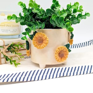 May include: A cream ceramic plant pot decorated with three yellow sunflowers and green leaves, holding a green plant. The pot sits on a white surface with a blue and white striped ribbon.