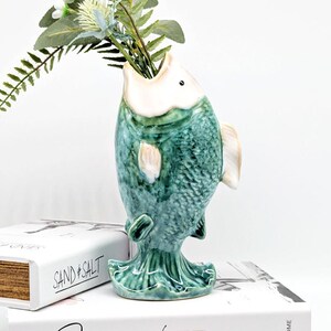 May include: A ceramic fish-shaped vase in shades of teal and cream, holding greenery. The vase has a textured scale pattern and is perched on a stack of books. The fish's mouth is open, serving as the vase opening.