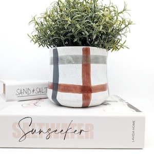 May include: A white ceramic planter with a red, grey, and blue plaid pattern. The planter is filled with a green plant and is sitting on a stack of books. The top book has the text "Sunseeker" printed on it.