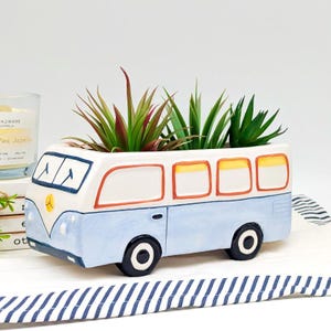 Puede incluir: Una maceta de cerámica con forma de autobús vintage, pintada en azul claro, blanco y naranja. El autobús tiene ruedas y ventanas negras, y está lleno de suculentas falsas verdes y rojas. Un artículo decorativo para la decoración del hogar.