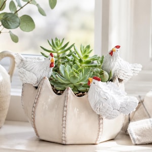 May include: A cream-coloured ceramic planter with a scalloped rim, featuring three white chicken figurines with red combs and yellow beaks attached to the exterior. The planter is filled with green succulent plants, showcasing a textured finish.