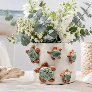 May include: A cylindrical white ceramic planter decorated with five raised, three-dimensional sea turtle figures. Each turtle features a textured, light green shell and brown flippers. The planter is filled with a bouquet of white flowers.